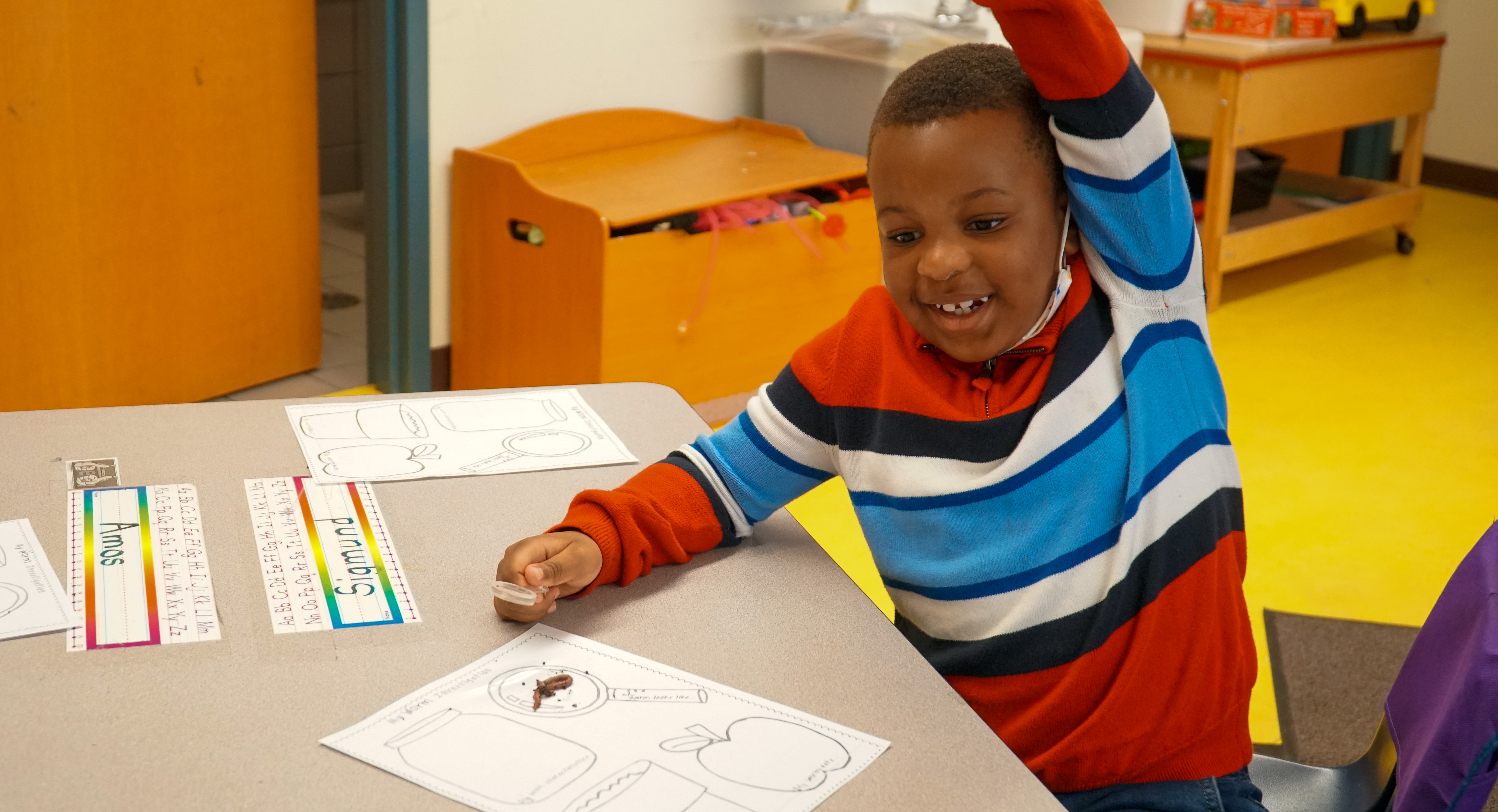 A boy with a worm smiles while having his hand up. 