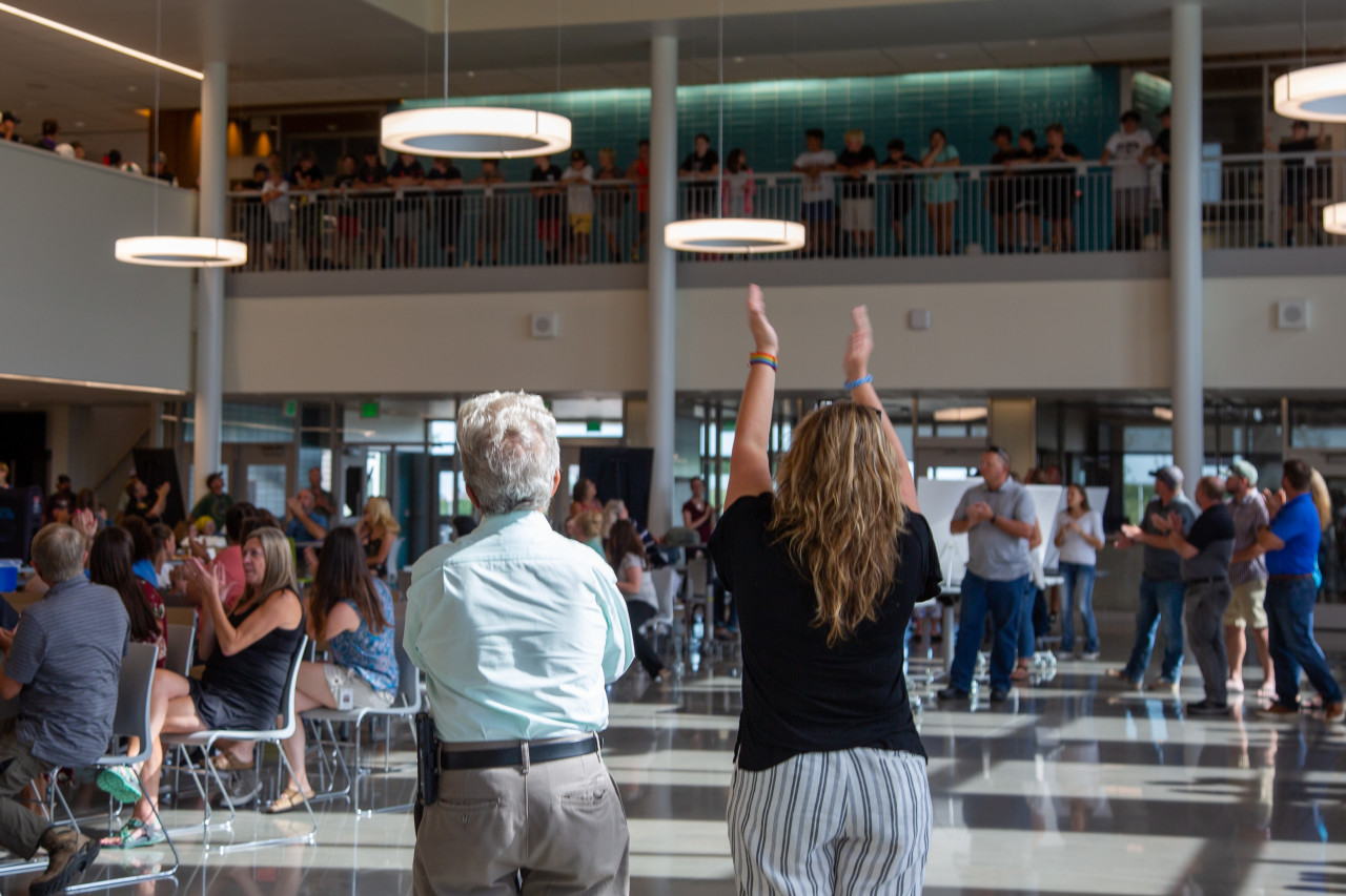 People in the common space look up to the balcony, clapping to those standing there. 