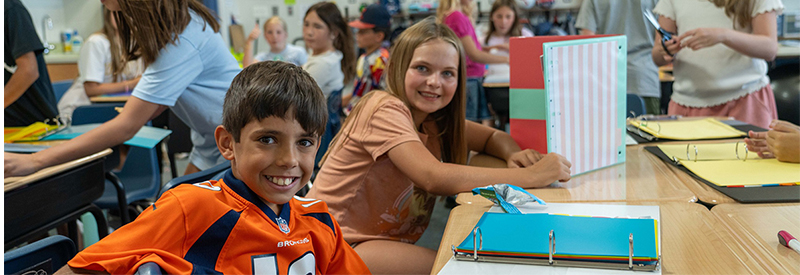 Two middle school students in a classroom with other kids.