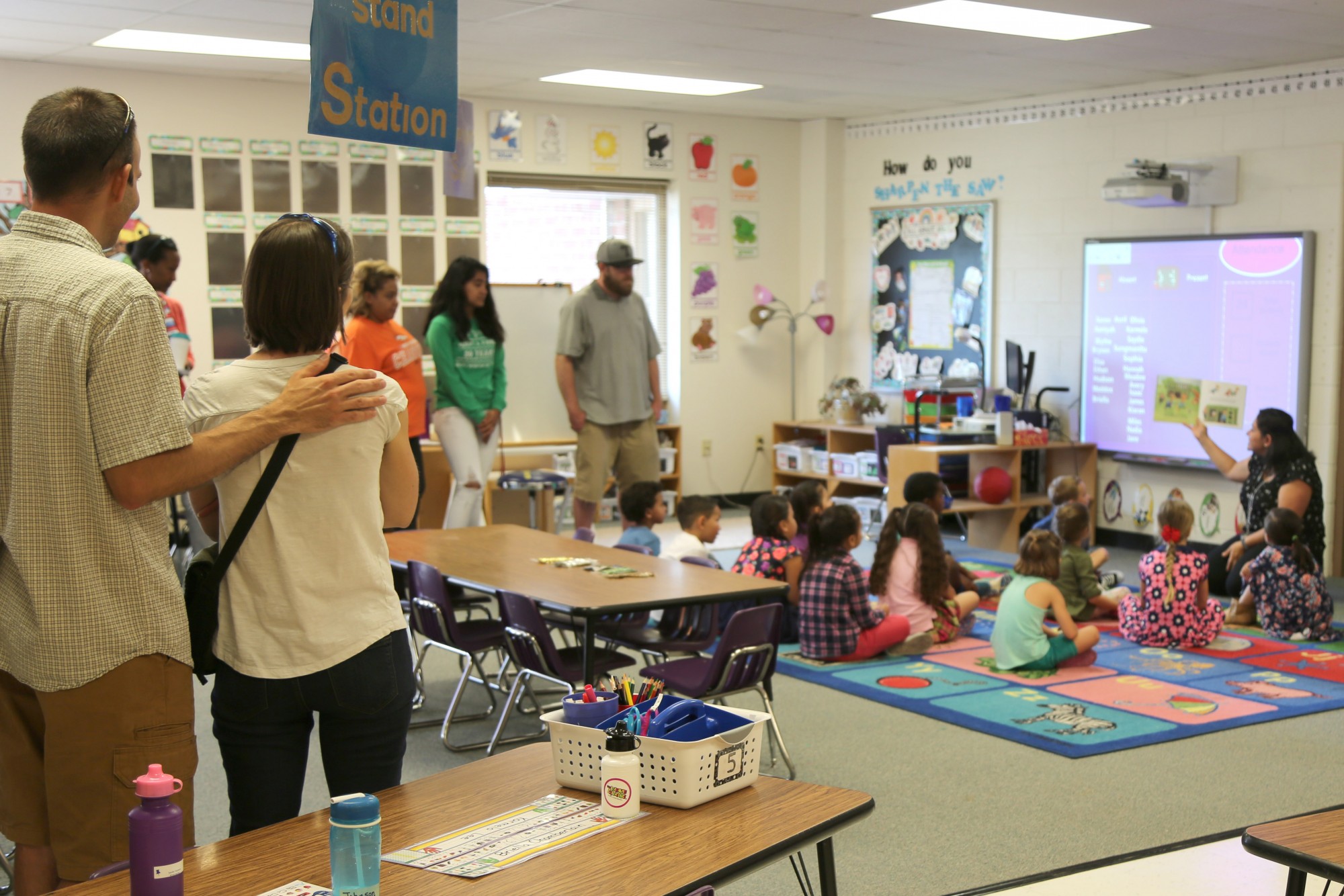A teacher reads to kindergarten students sitting on a rug while parents watch.