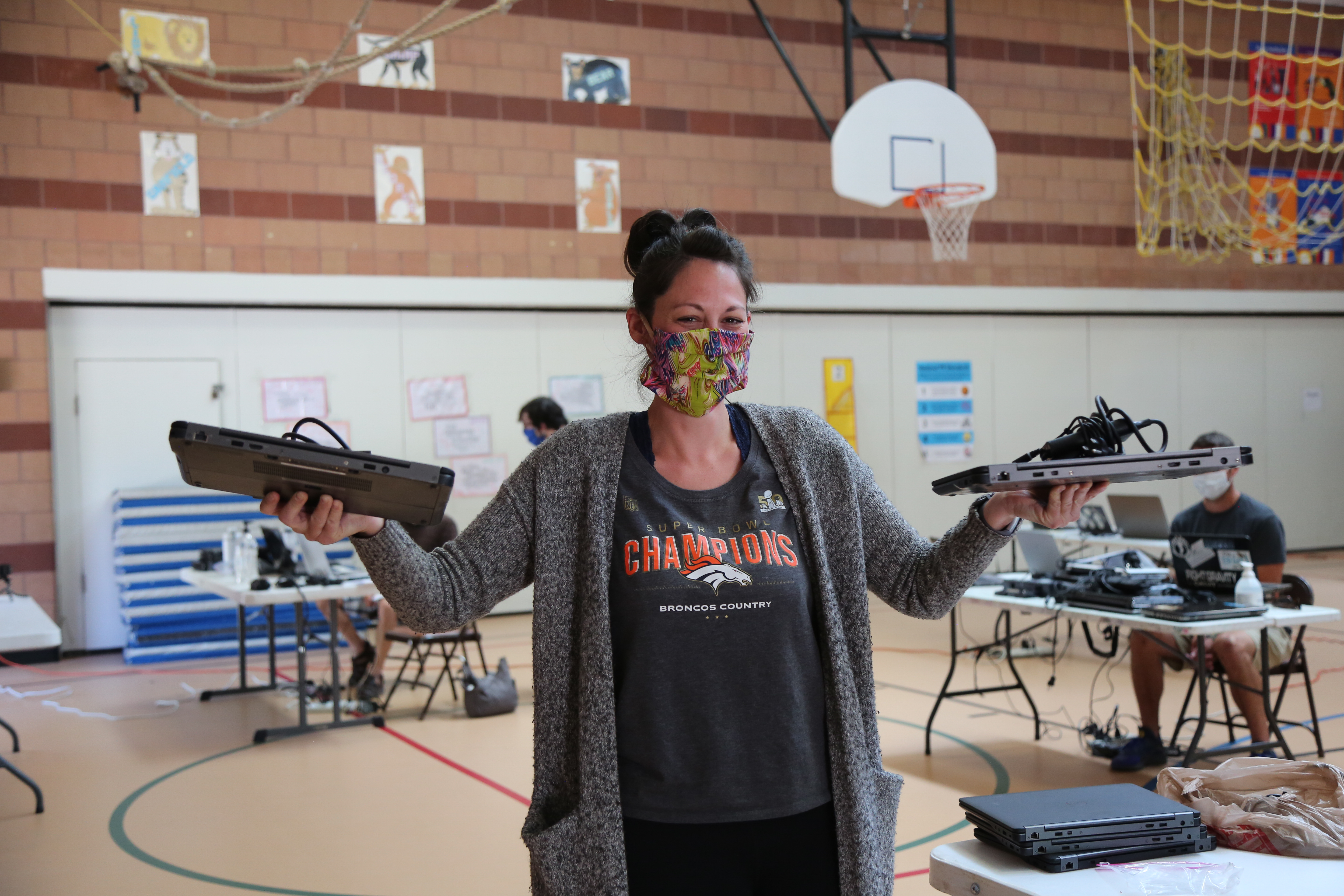 A teacher wearing a face mask enthusiastically holds up a donated laptop in each hand.