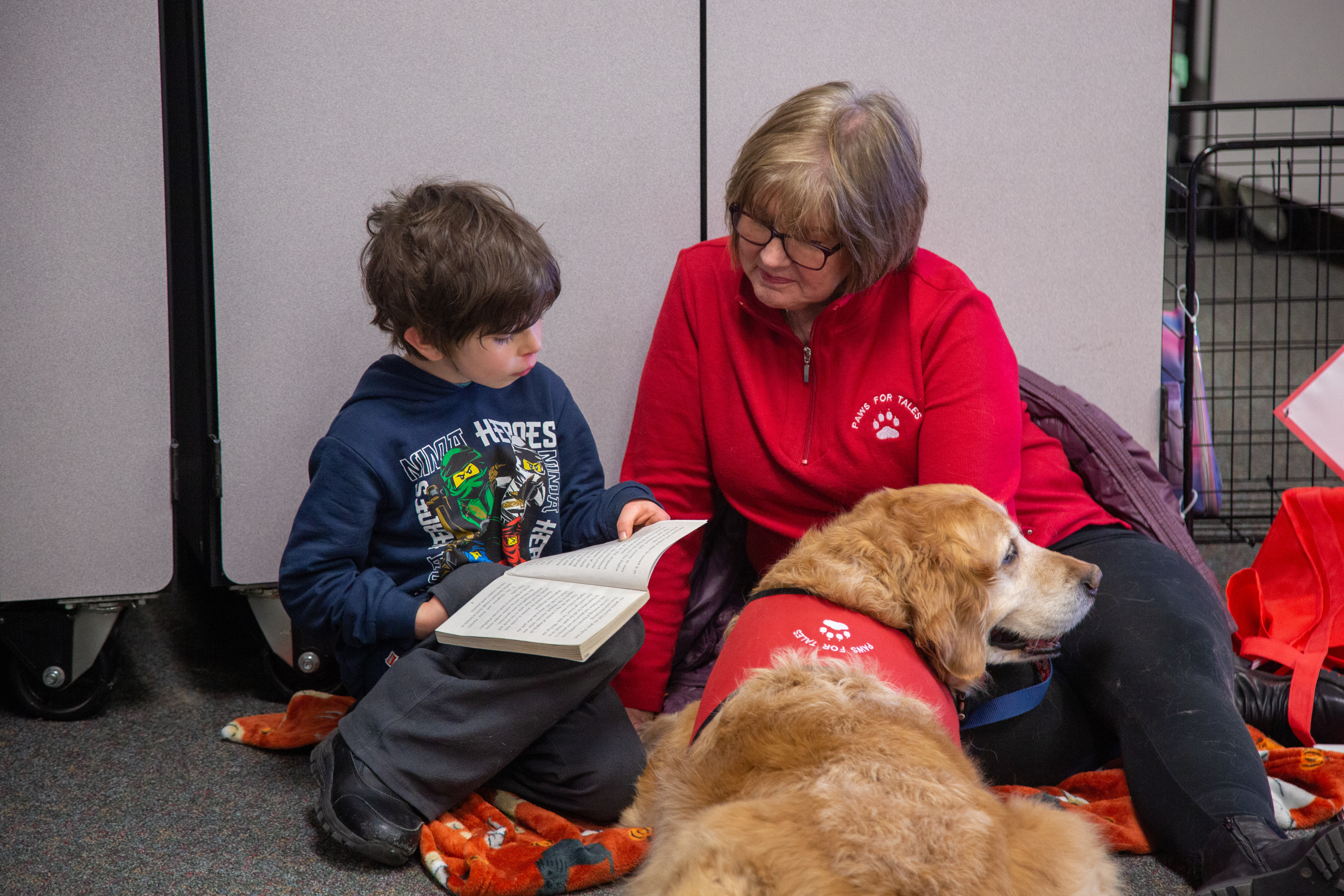 A young student reads to a dog and a volunteer. 