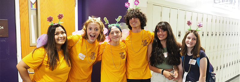 High school students wearing fun head pieces stand in front of lockers, smiling at the camera. 