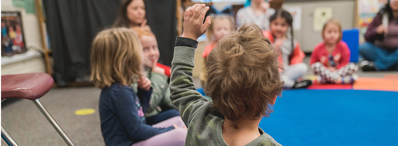 Kindergartners sit on a rug in a classroom. 