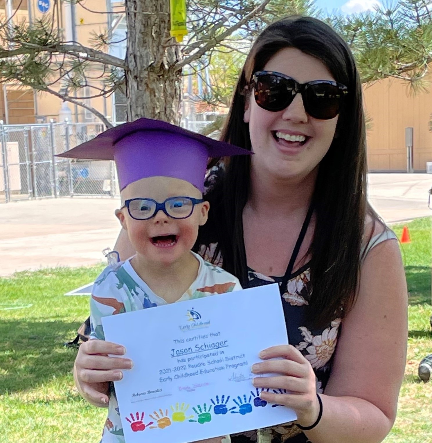 Little boy wearing a graduation cap standing in front of a woman holding his graduation certificate