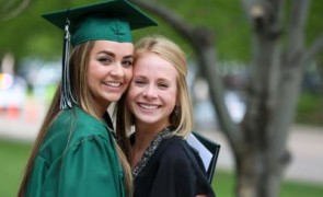 A graduate in cap and gown stands with a friend smiling.